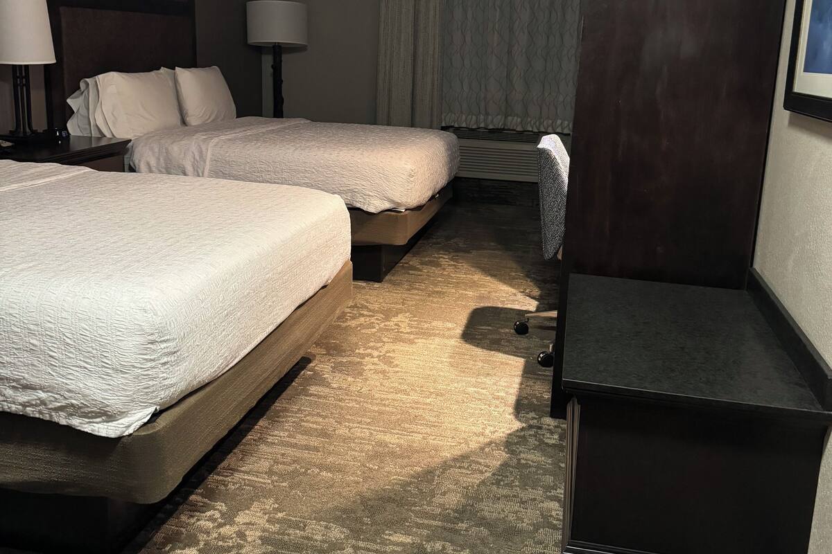 Quarto de hotel com duas camas, uma escrivaninha, uma cadeira, um guarda-roupa e uma janela com cortinas.