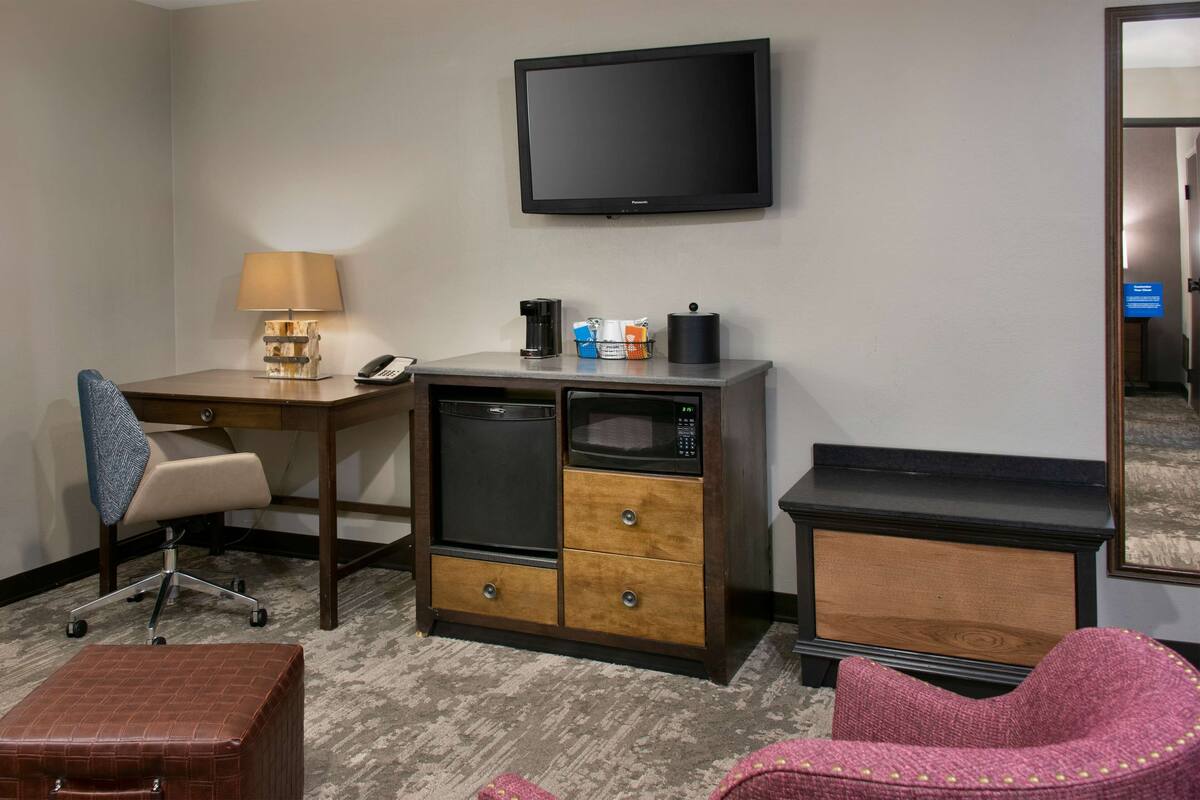 Quarto de hotel com escrivaninha, cadeira, micro-ondas, máquina de café e televisão na parede.
