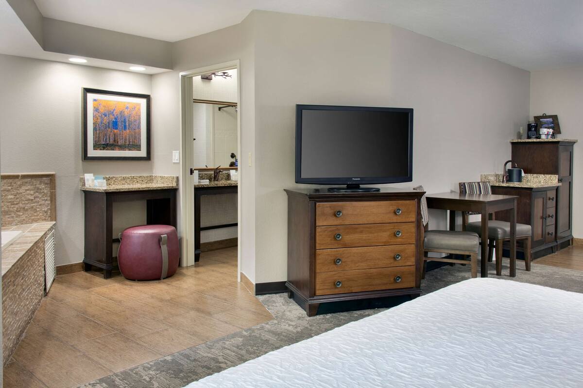 Quarto de hotel com cama, televisão, penteadeira, escrivaninha e banheiro visível através de uma porta aberta.