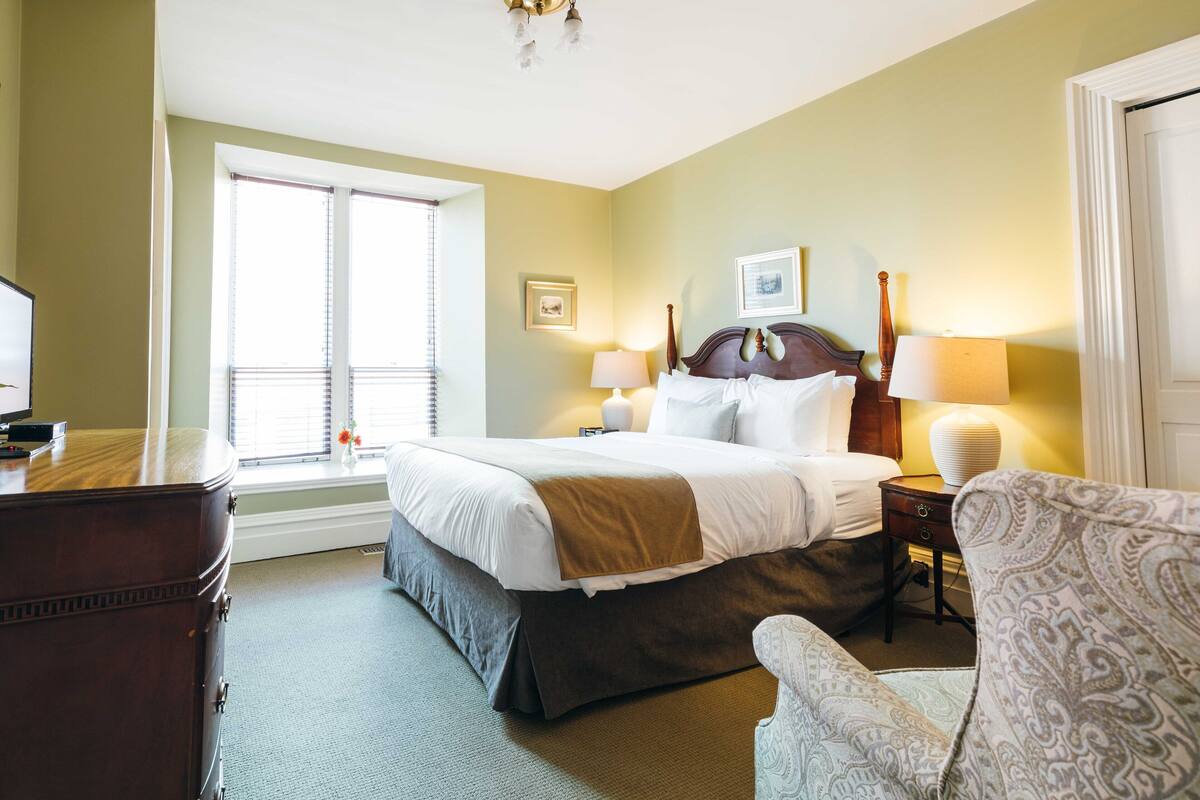 A hotel room with a large bed, two bedside lamps, a dresser, a chair, and a window with blinds.