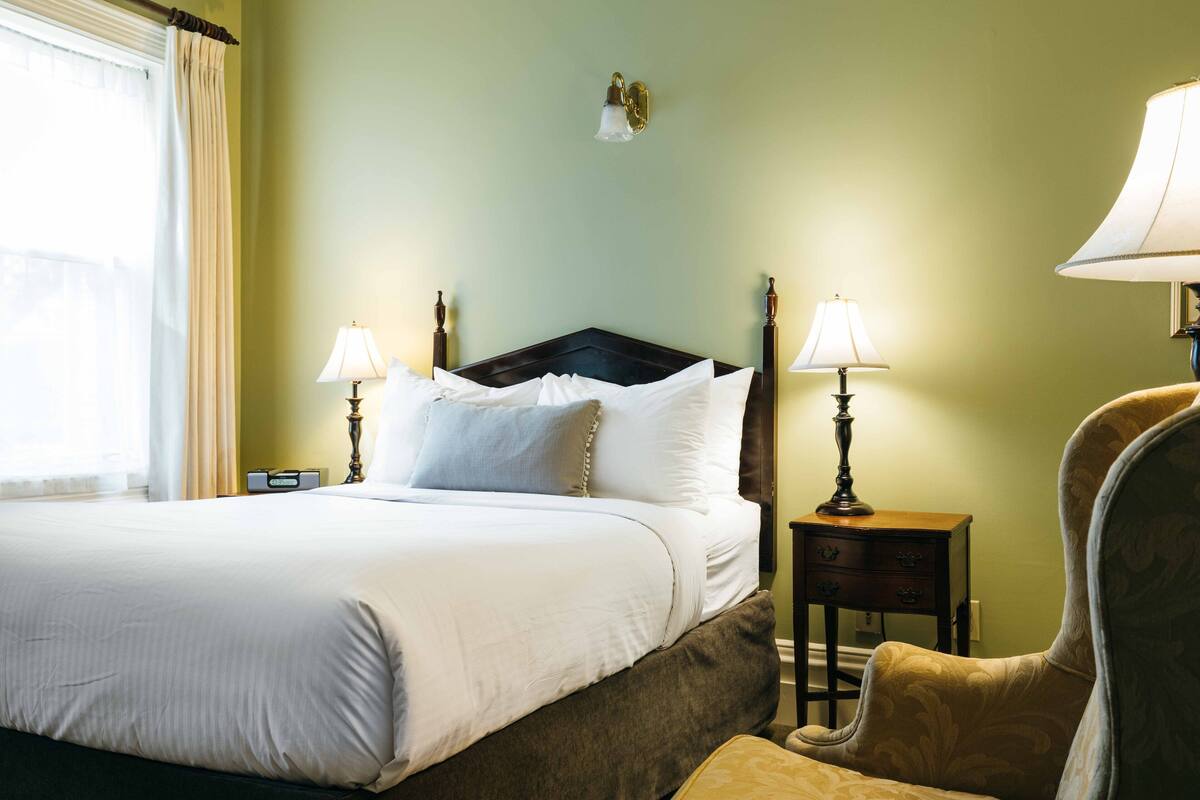 A neatly made bed with white linens, two bedside lamps, a nightstand, and a chair.