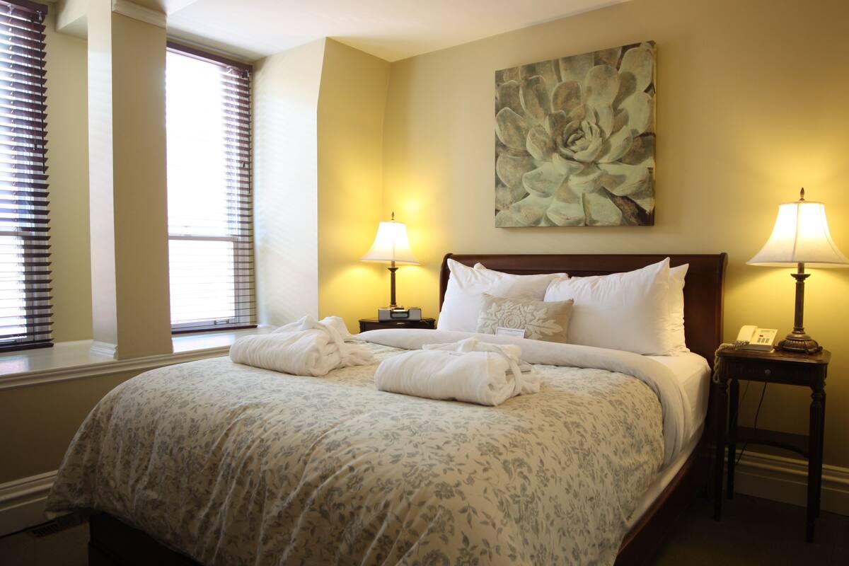 A neatly made bed with a floral bedspread, two bedside lamps, a painting above the bed, and a window with blinds.