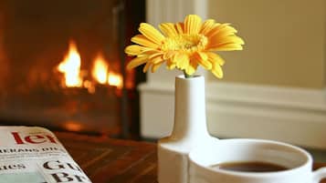 A newspaper, a cup of coffee, a spoon, and a vase with a yellow flower on a wooden table with a fireplace in the background.