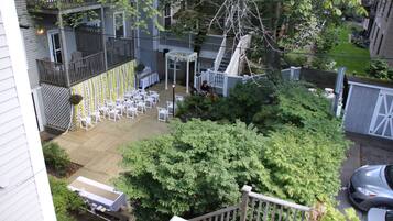 A wooden deck with white chairs and a covered seating area, surrounded by greenery and a fence.