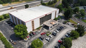 Une vue aérienne d’un grand bâtiment doté d’un toit plat, d’un parking avec plusieurs voitures et d’une route en arrière-plan.