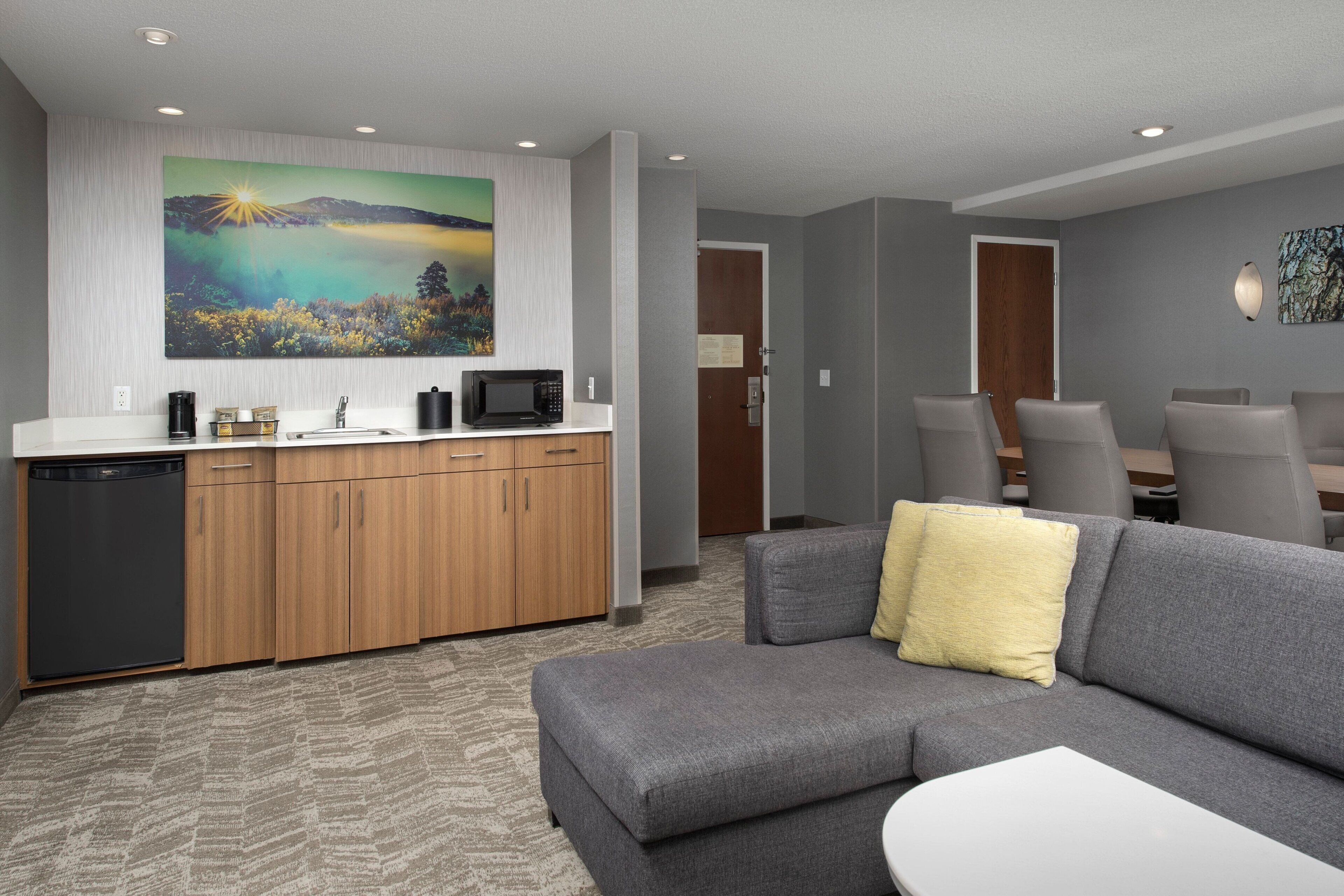 A modern living room with a gray sofa, a small white table, and a gray couch with a yellow pillow. There is a kitchen area with wooden cabinets and a microwave.