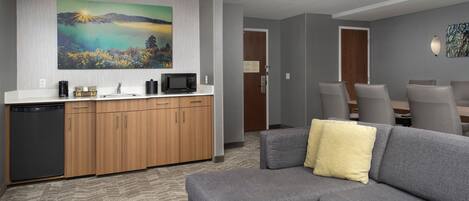 Un salón moderno con un sofá gris, una mesita blanca y un sofá gris con un cojín amarillo. Se observa una zona de cocina con armarios de madera y un microondas.