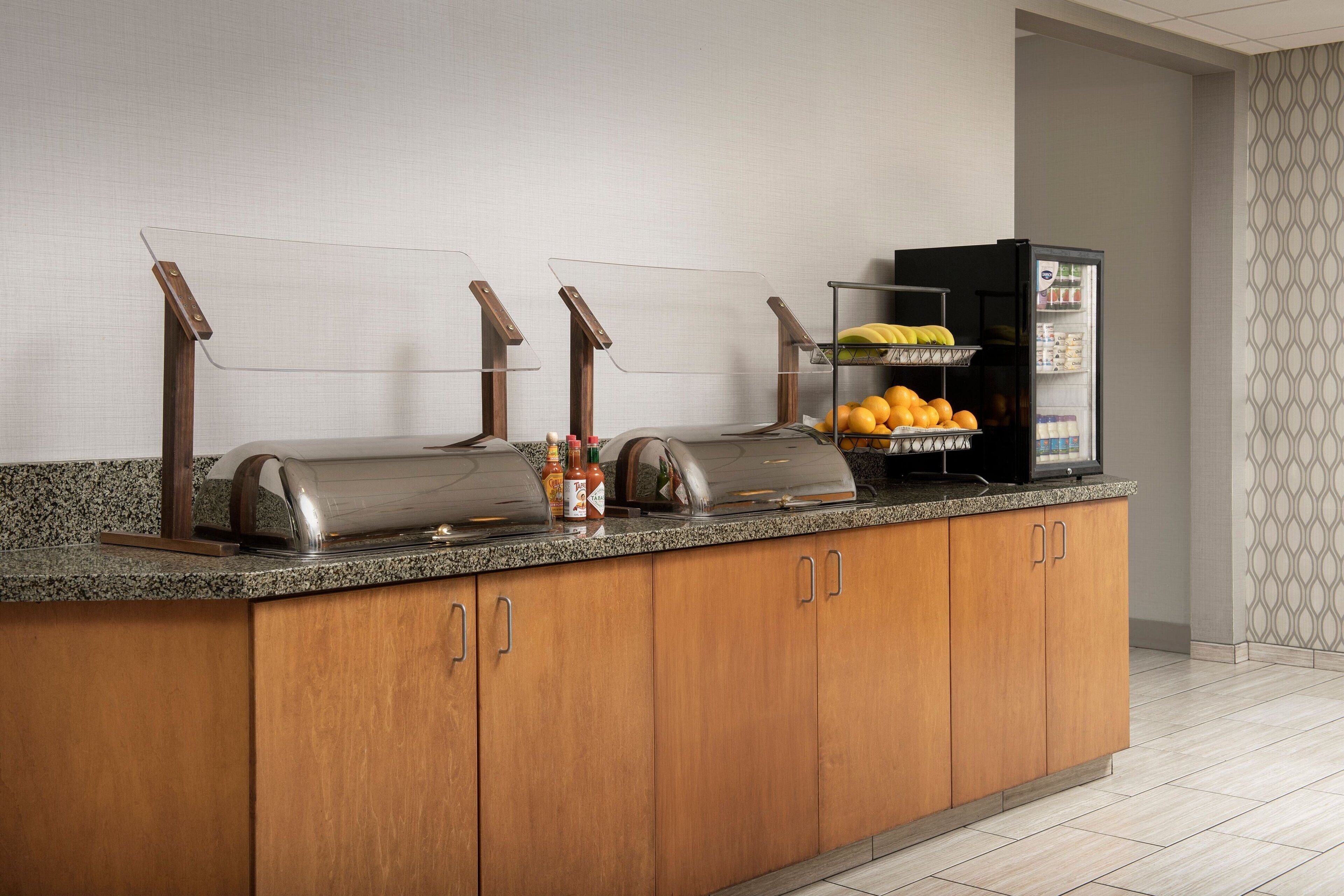 A buffet setup with a refrigerator, condiment station, and a fruit display.