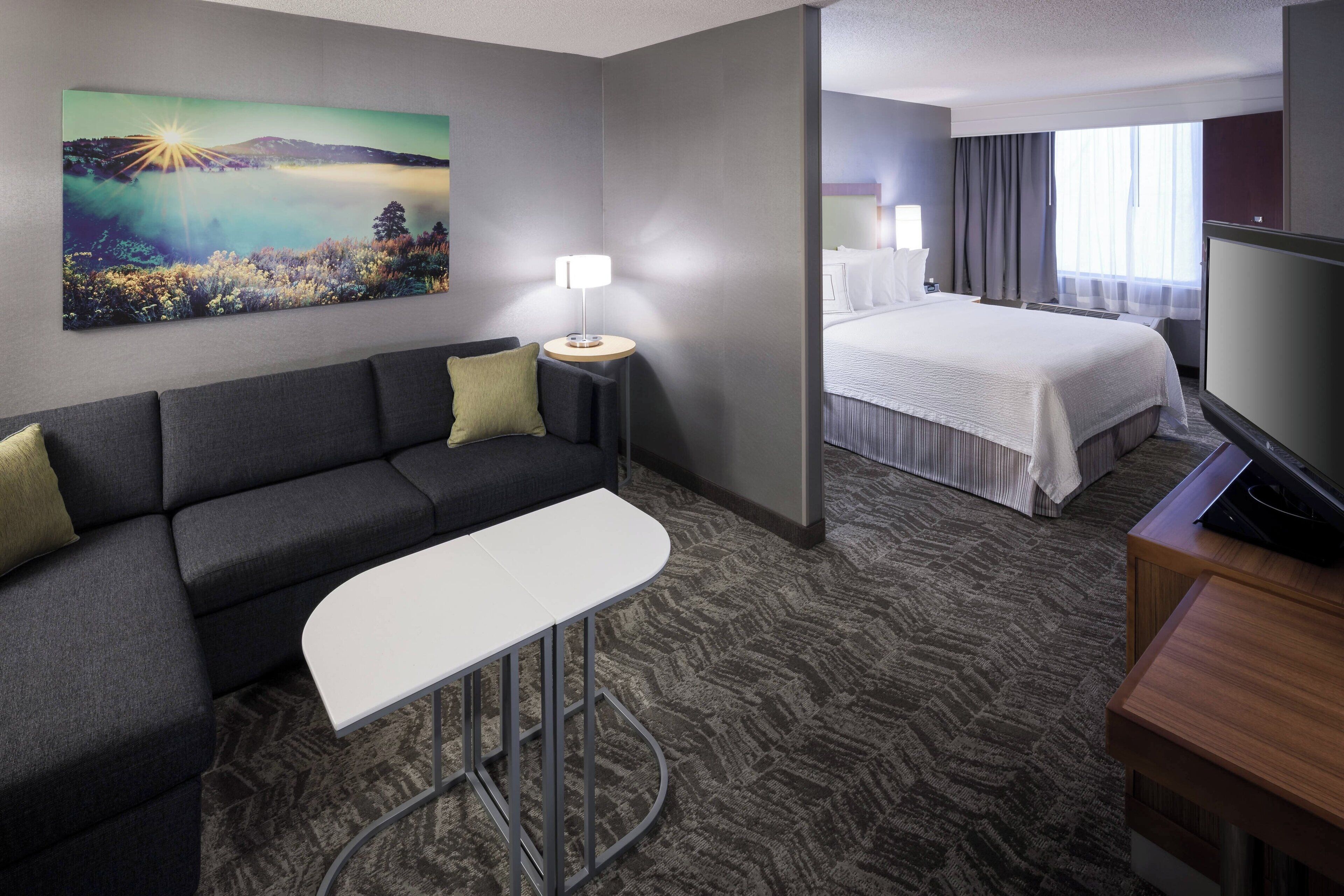 A hotel room with a sofa, a small table, a bed, a TV, and a painting on the wall.