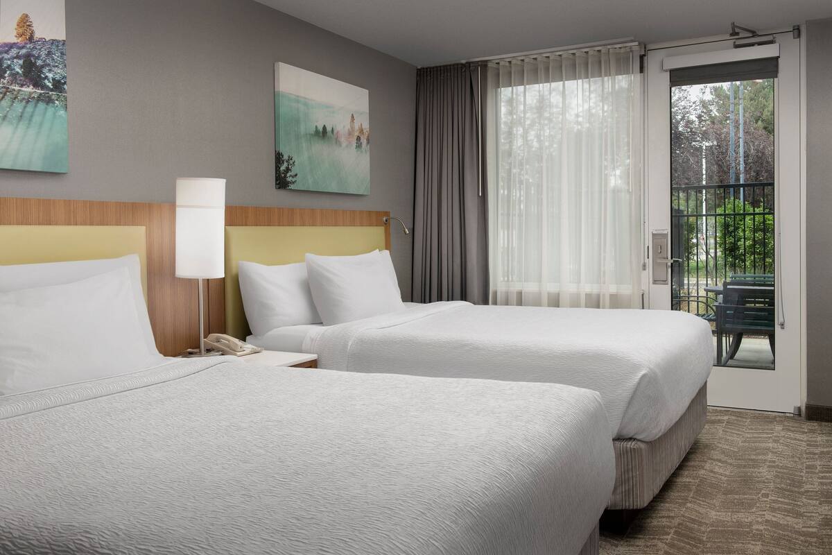 A hotel room with two beds, a nightstand with a lamp, and a view of the outdoors through a door.