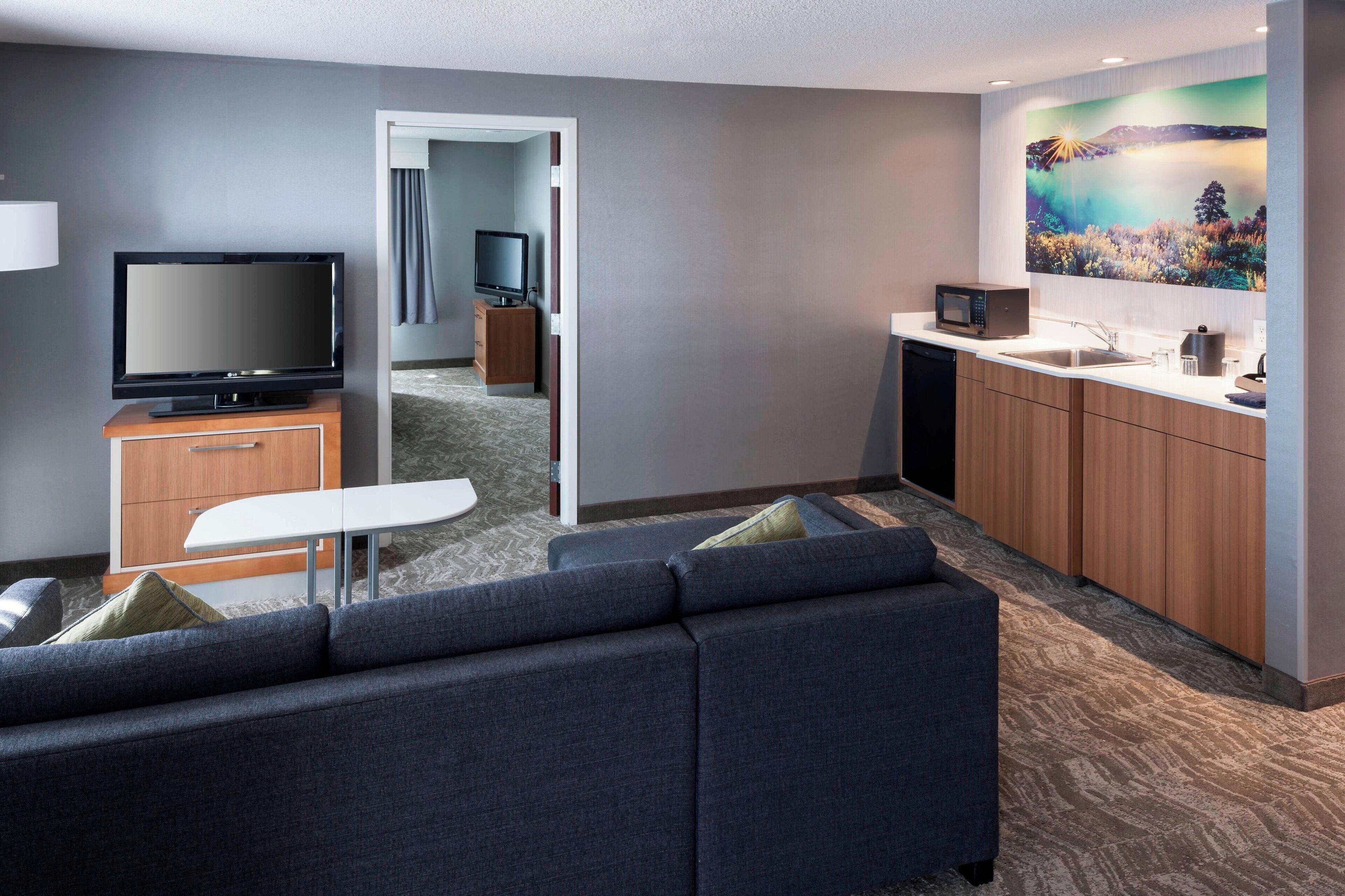 A modern hotel room with a sofa, TV, kitchenette, and a painting on the wall.