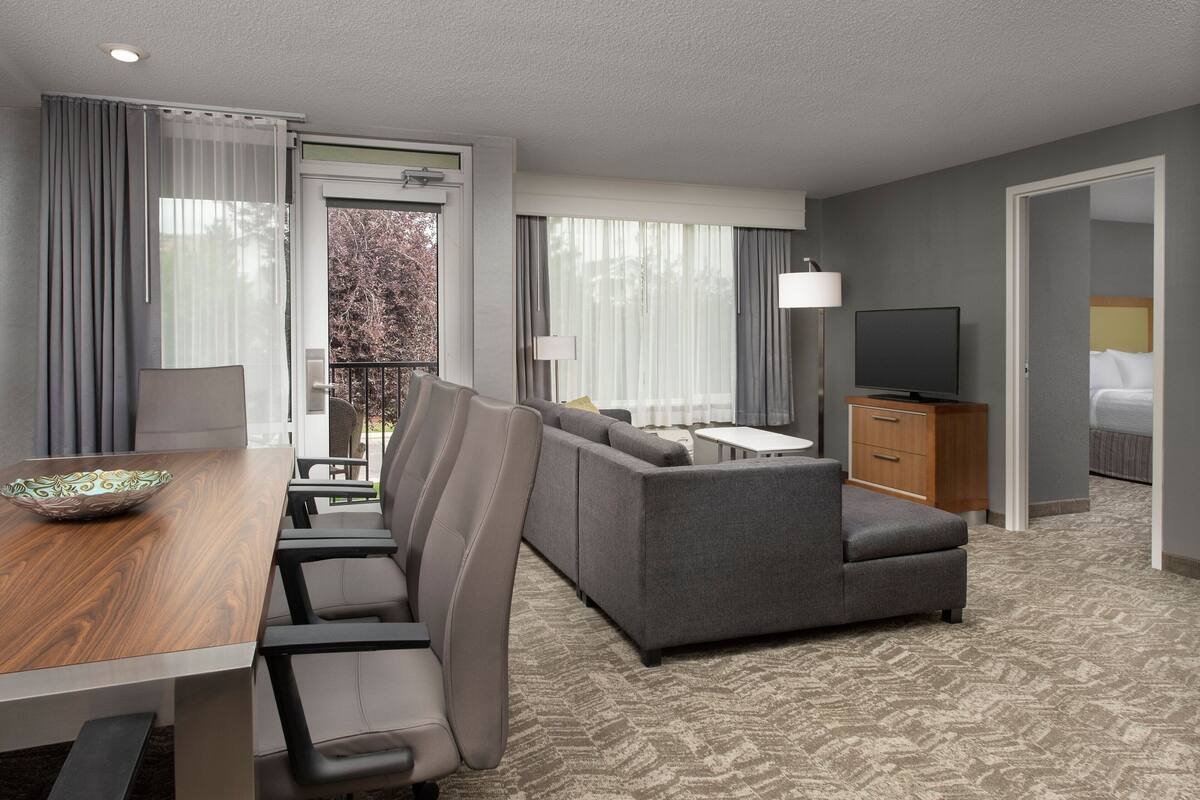 A hotel room with a dining area, a sofa, a TV, and a balcony with curtains.
