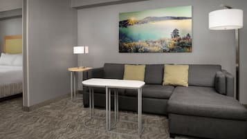A hotel room with a bed, a gray sofa, a small table, and a painting of a landscape on the wall.