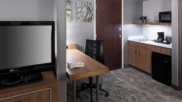 A hotel room with a TV, a wooden desk, a chair, a door, and a kitchenette.