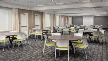 A conference room with multiple tables and chairs, carpeted flooring, and large windows with curtains.