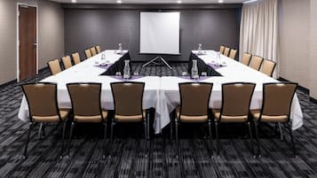 Una sala de conferencias con una larga mesa, sillas, una pantalla de proyector y piso alfombrado.