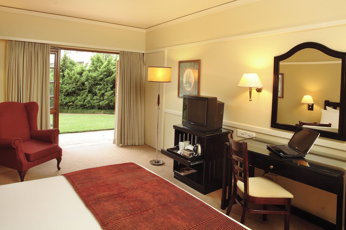 Una habitación de hotel con una cama, un escritorio con una computadora portátil, un televisor, una silla y un ventanal con cortinas.