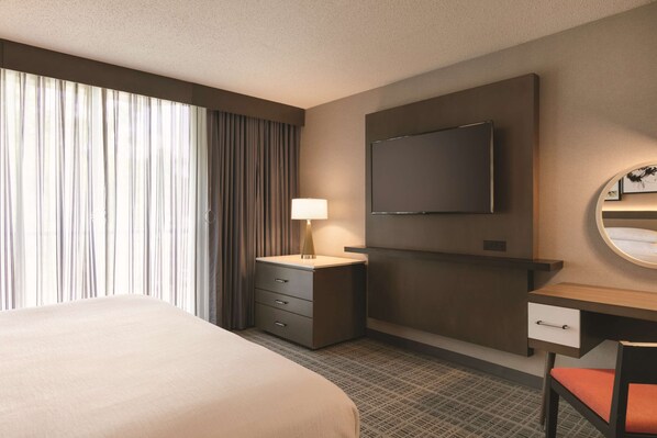 A hotel room with a bed, a desk, a chair, a TV, a lamp, and a window with curtains.