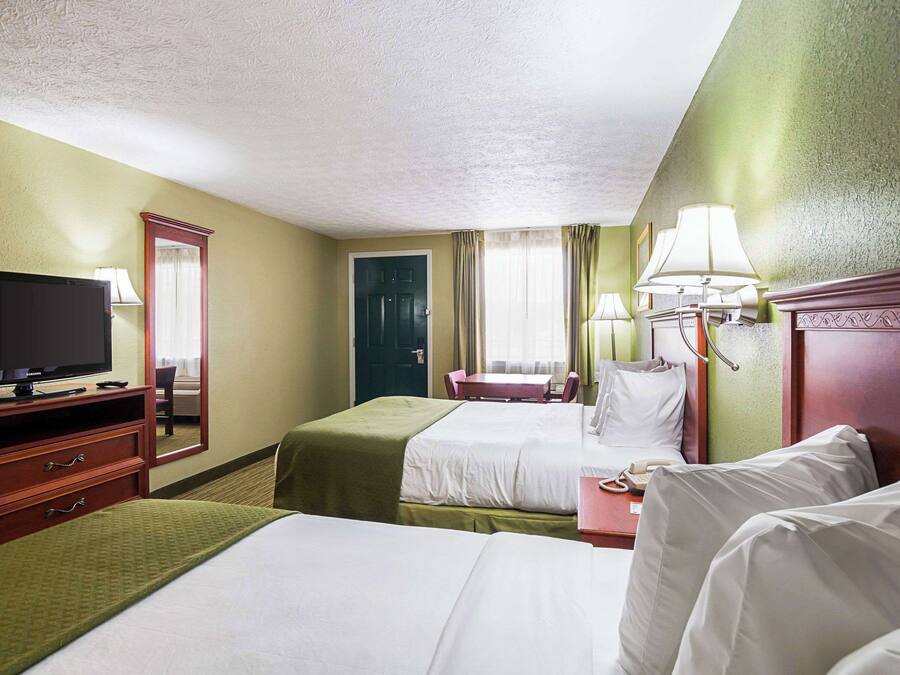 A hotel room with two beds, a television, a desk with a lamp, and a door.