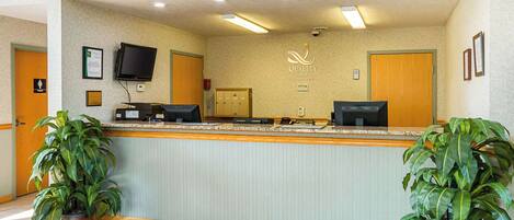 A hotel reception area with a reception desk, two potted plants, a television, and a sign that reads "Quality Inn."