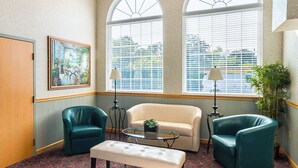 Une pièce avec un tapis à motifs floraux, un canapé beige, deux fauteuils turquoise, une table basse en verre, un banc, un tableau et une grande fenêtre avec des stores.