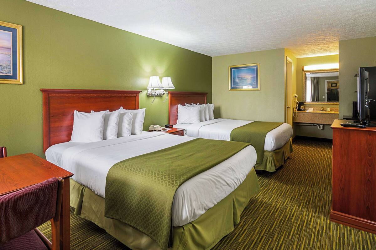 A hotel room with two beds, a desk, a chair, a TV, and a bathroom with a mirror and sink.