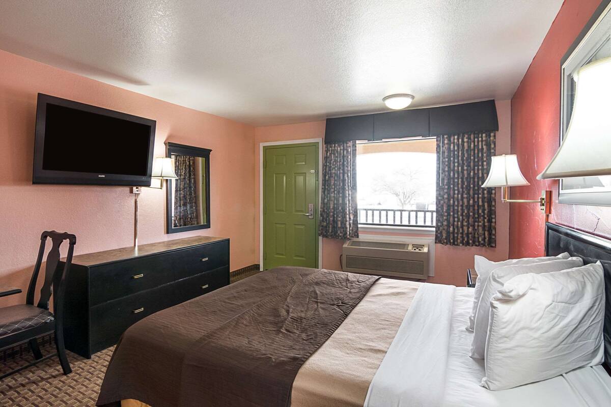 A hotel room with a bed, a desk with a chair, a television mounted on the wall, a green door, and a window with curtains.