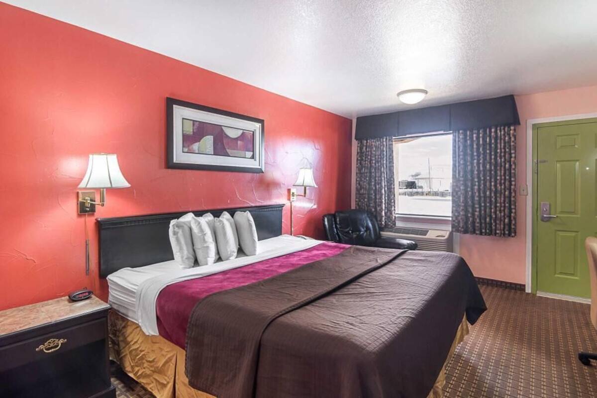 A hotel room with a bed, two bedside lamps, a framed picture on the wall, a green door, and a window with curtains.