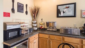 A kitchenette with a microwave, toaster, and coffee maker. There are various food containers and a fruit basket on the counter.