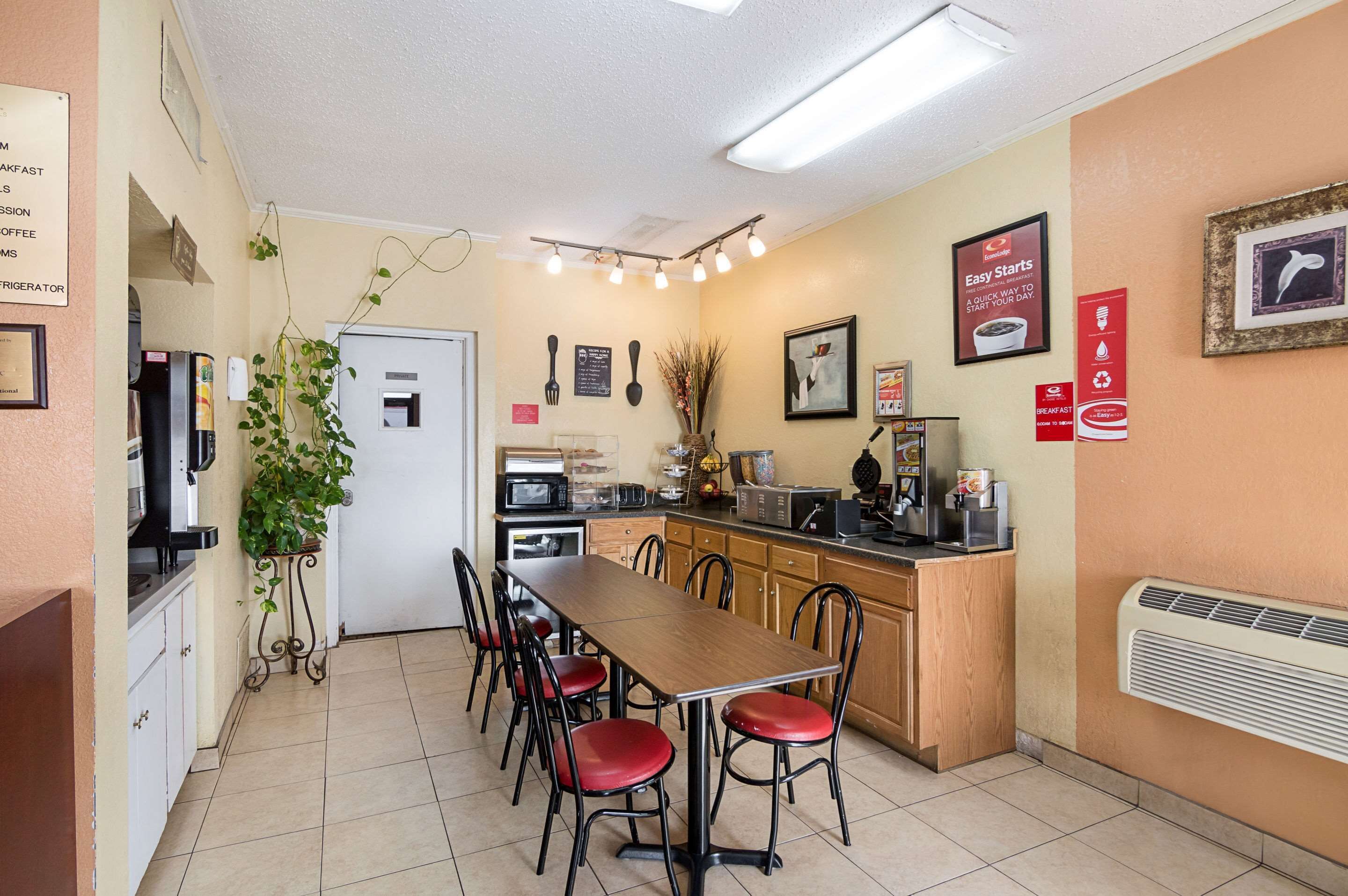 Cocina pequeña y bien iluminada con una mesa y sillas, un refrigerador, un microondas y un lavamanos. Hay letreros en la pared y una planta en una esquina.