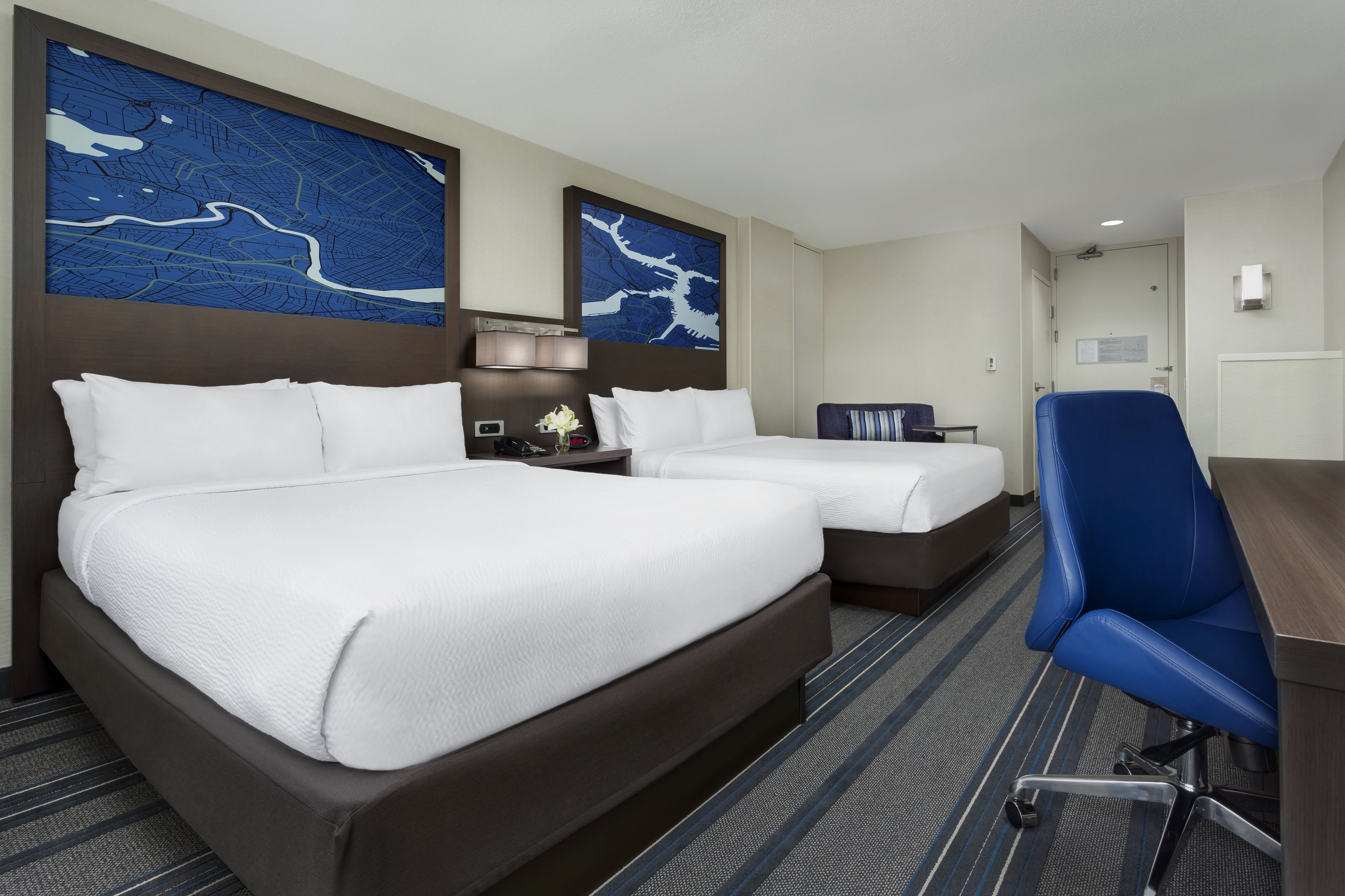 A hotel room with two beds, a desk, and a chair. There are two large wall art pieces above the beds.