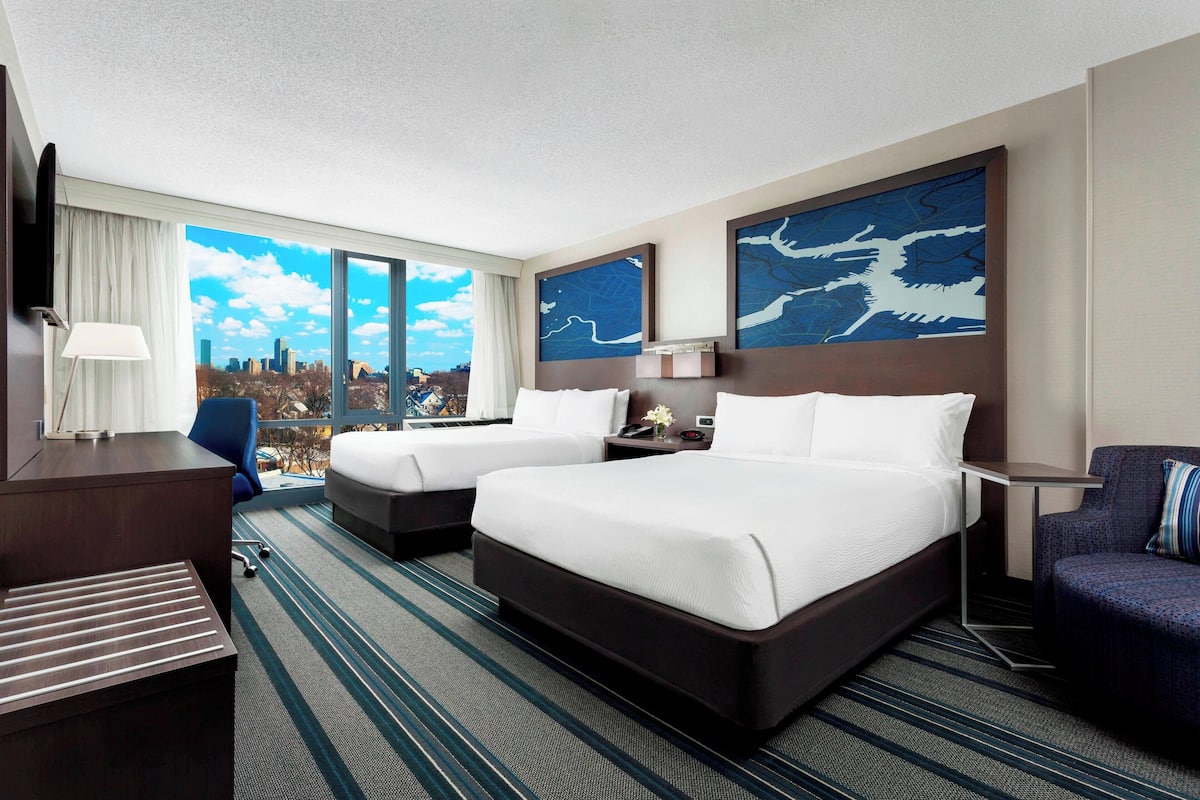 Quarto de hotel com duas camas, uma escrivaninha, uma cadeira e grandes janelas com vista para a cidade.