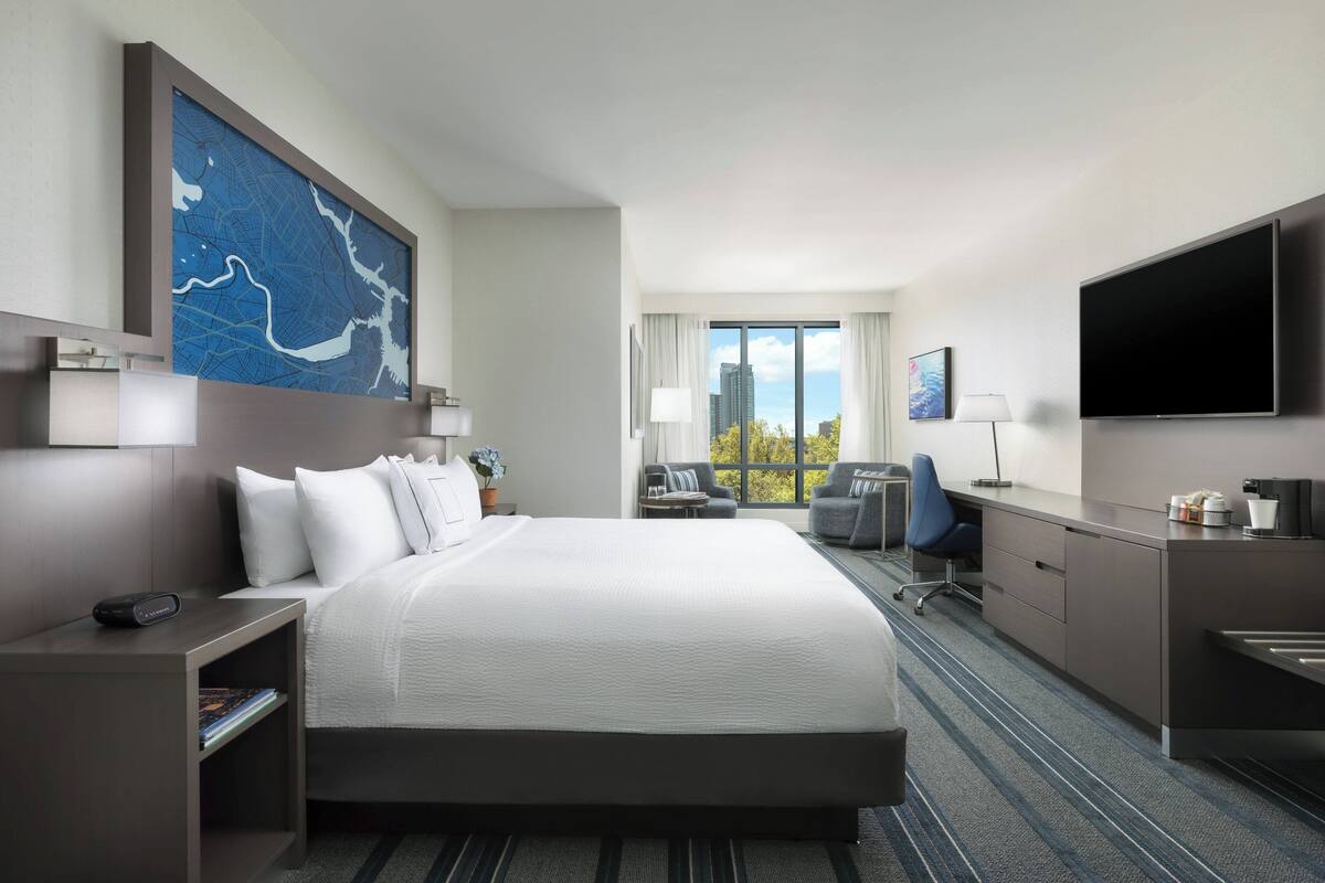 Quarto de hotel com uma cama grande, uma escrivaninha com cadeira, televisão de tela plana, uma obra de arte com um mapa na parede e uma janela com vista para a cidade.