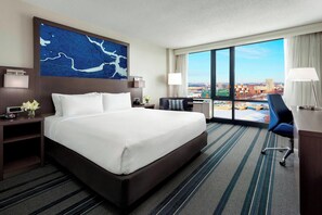 A hotel room with a large bed, a desk, a chair, and a view of the city. - Courtyard by Marriott Boston-Cambridge (Cambridge)