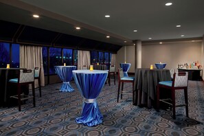 Una habitación de hotel con ventanas amplias, alfombra estampada y mesas con manteles azules.