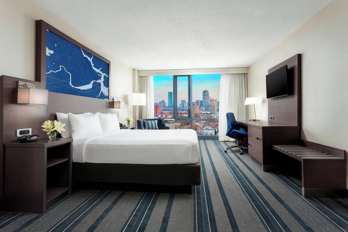 Quarto de hotel com uma cama grande, uma escrivaninha, televisão e vista para a cidade.