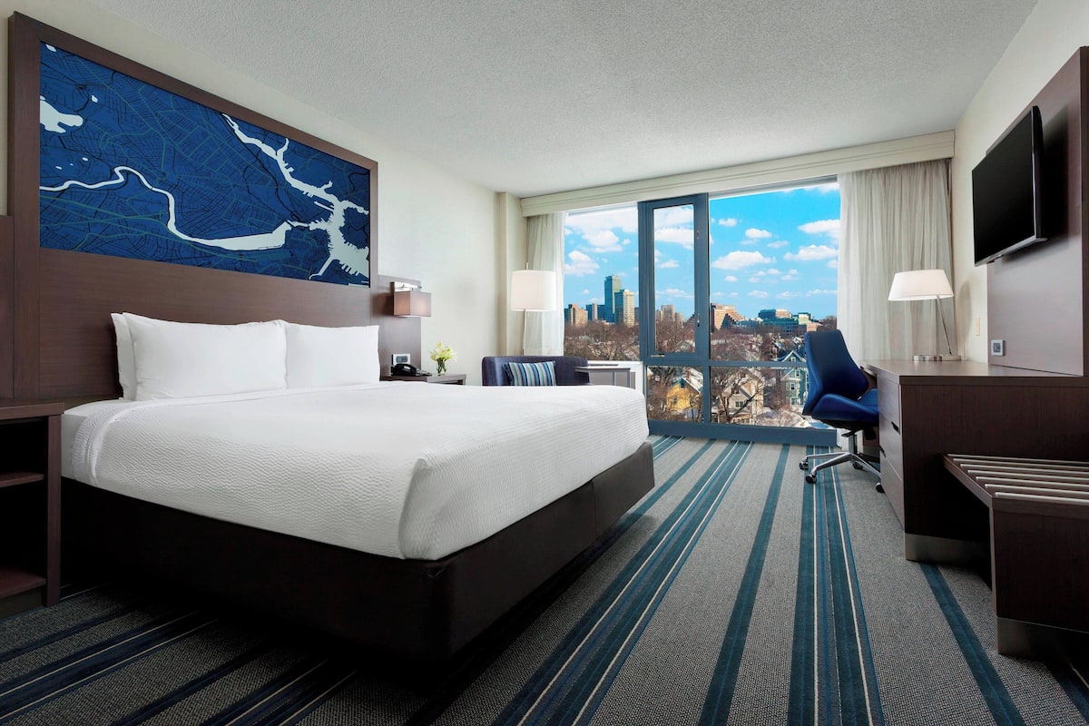 Quarto de hotel com uma cama grande, uma escrivaninha, uma cadeira, televisão e varanda com vista para a cidade.