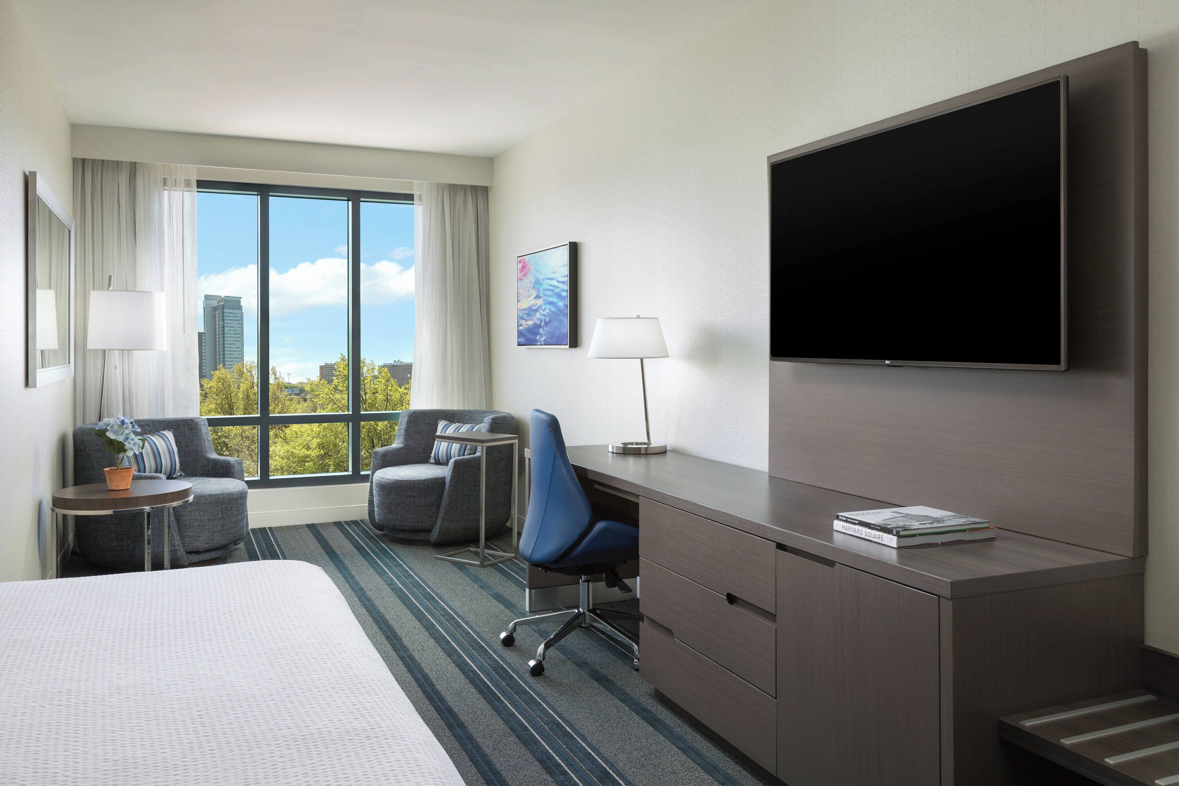 A hotel room with a large bed, a desk with a chair, a TV mounted on the wall, and a view of the city through large windows.