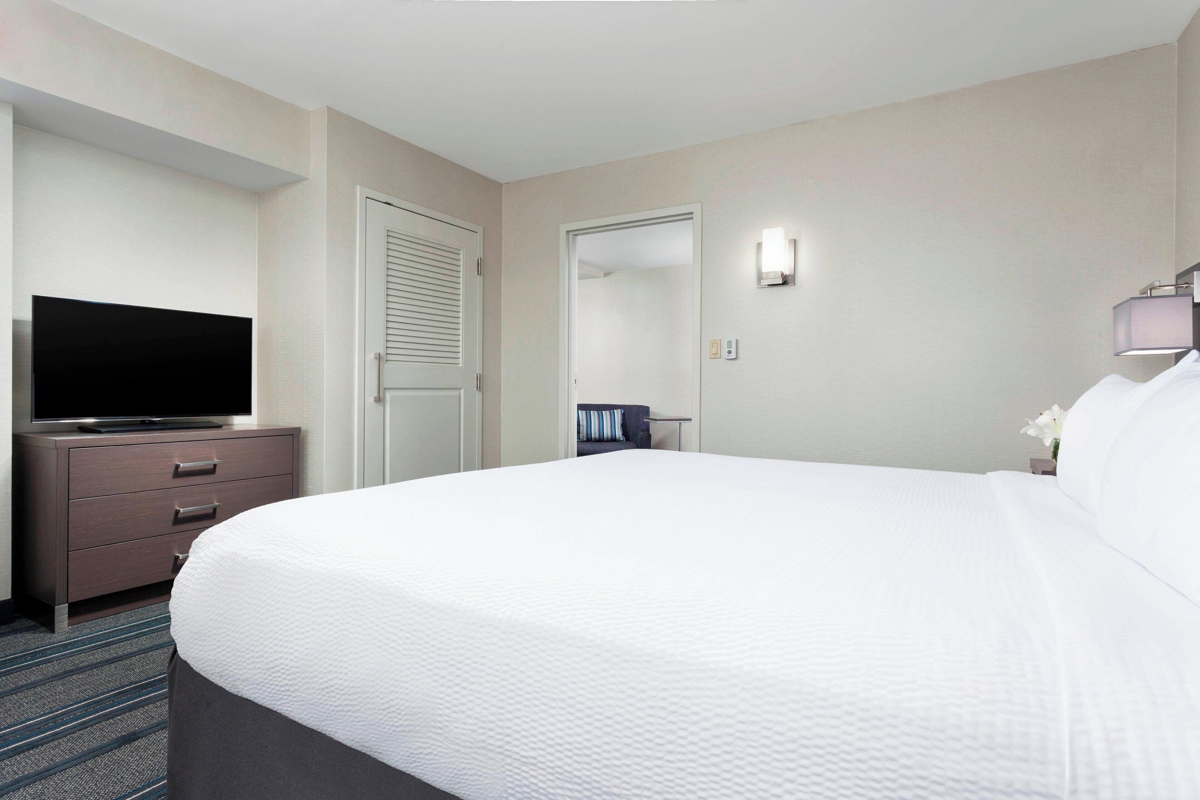 A hotel room with a large bed, a television on a dresser, a door, and a wall-mounted light.