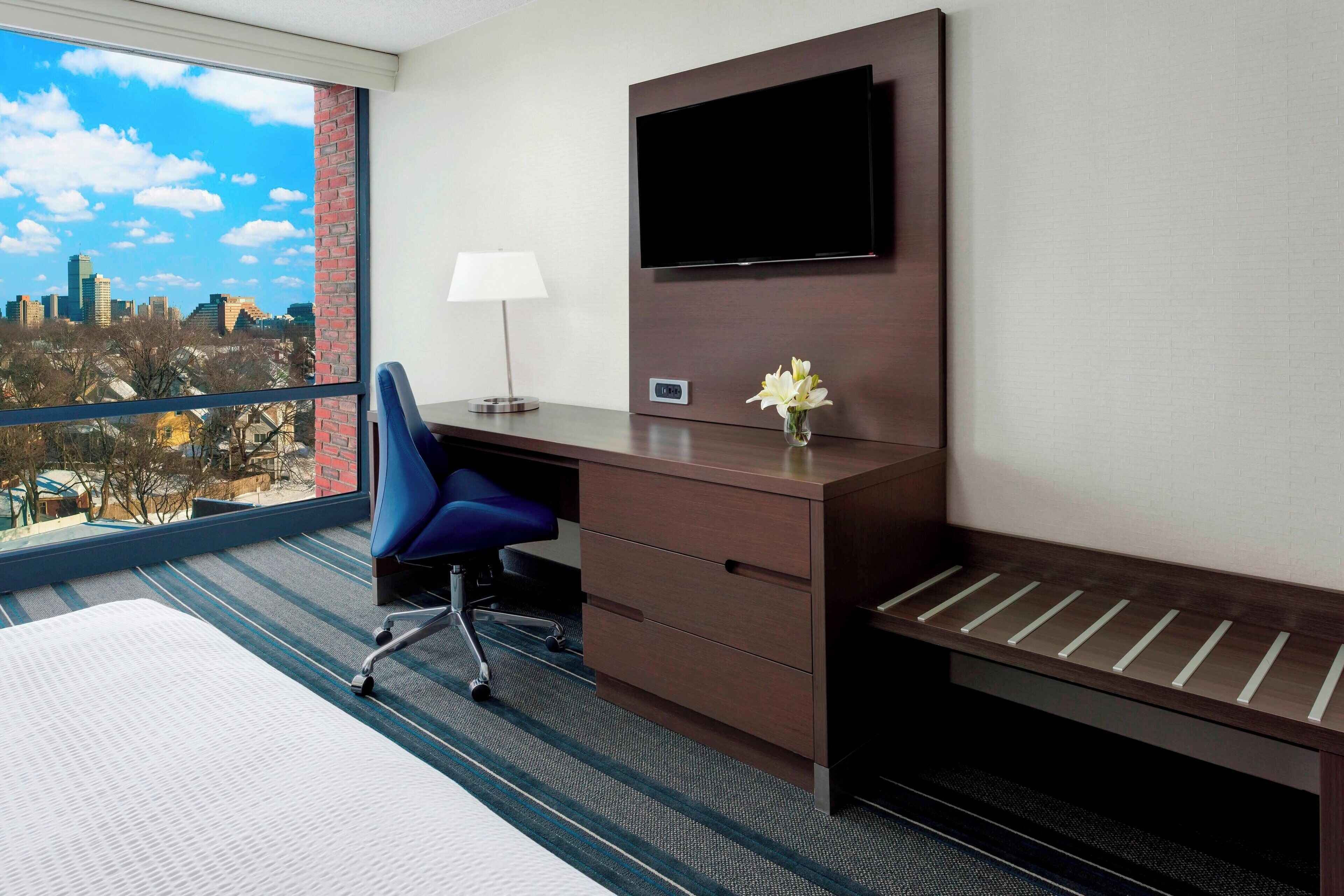A hotel room with a large window, a desk with a chair, a flat-screen TV mounted on the wall, and a small vase with flowers.