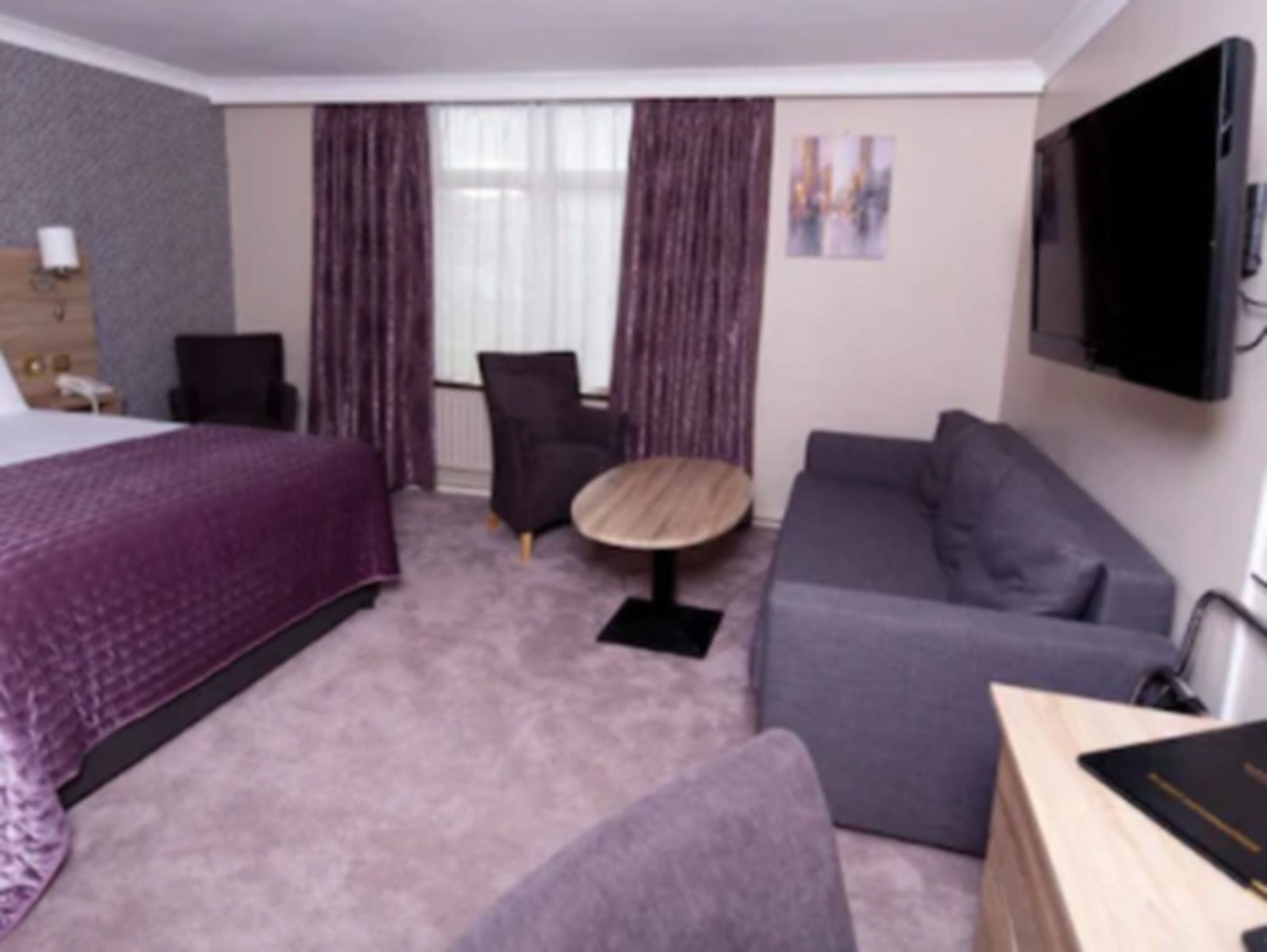 Una habitación de hotel con cama, un sofá gris, una mesa redonda, una silla, televisor montado en la pared y una ventana con cortinas.