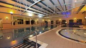 Piscina interna com água cristalina, chão ladrilhado e teto alto com vigas de metal.