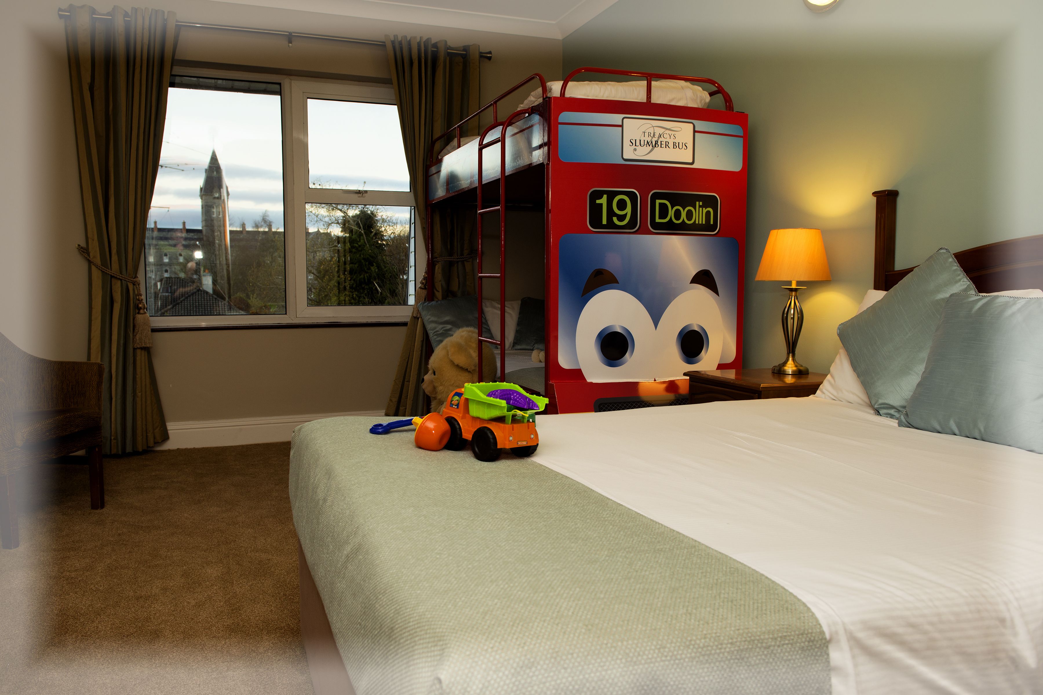 A hotel room with a bunk bed, a toy bus, a lamp, and a view of a cityscape through a window.