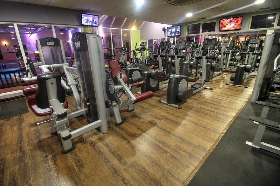 Uma academia com diversos equipamentos de exercícios, piso de madeira e várias telas de televisão.