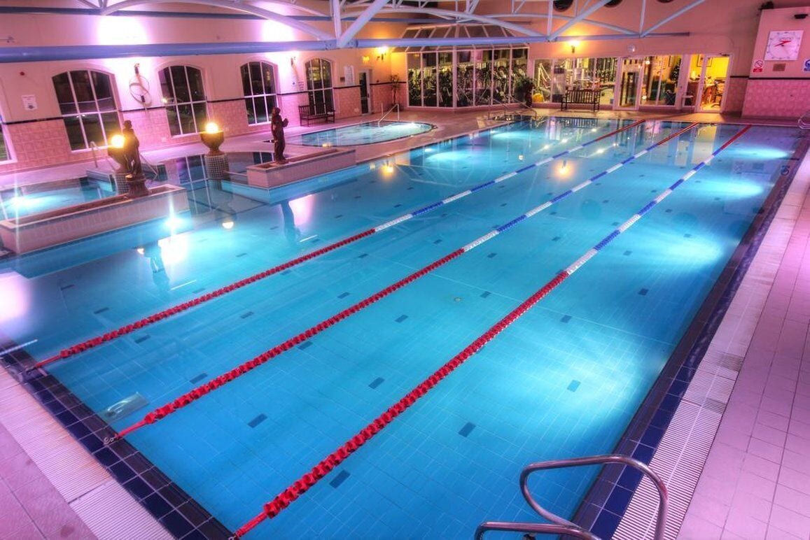 A large indoor swimming pool with multiple lanes, surrounded by a tiled floor and walls, and equipped with safety railings.