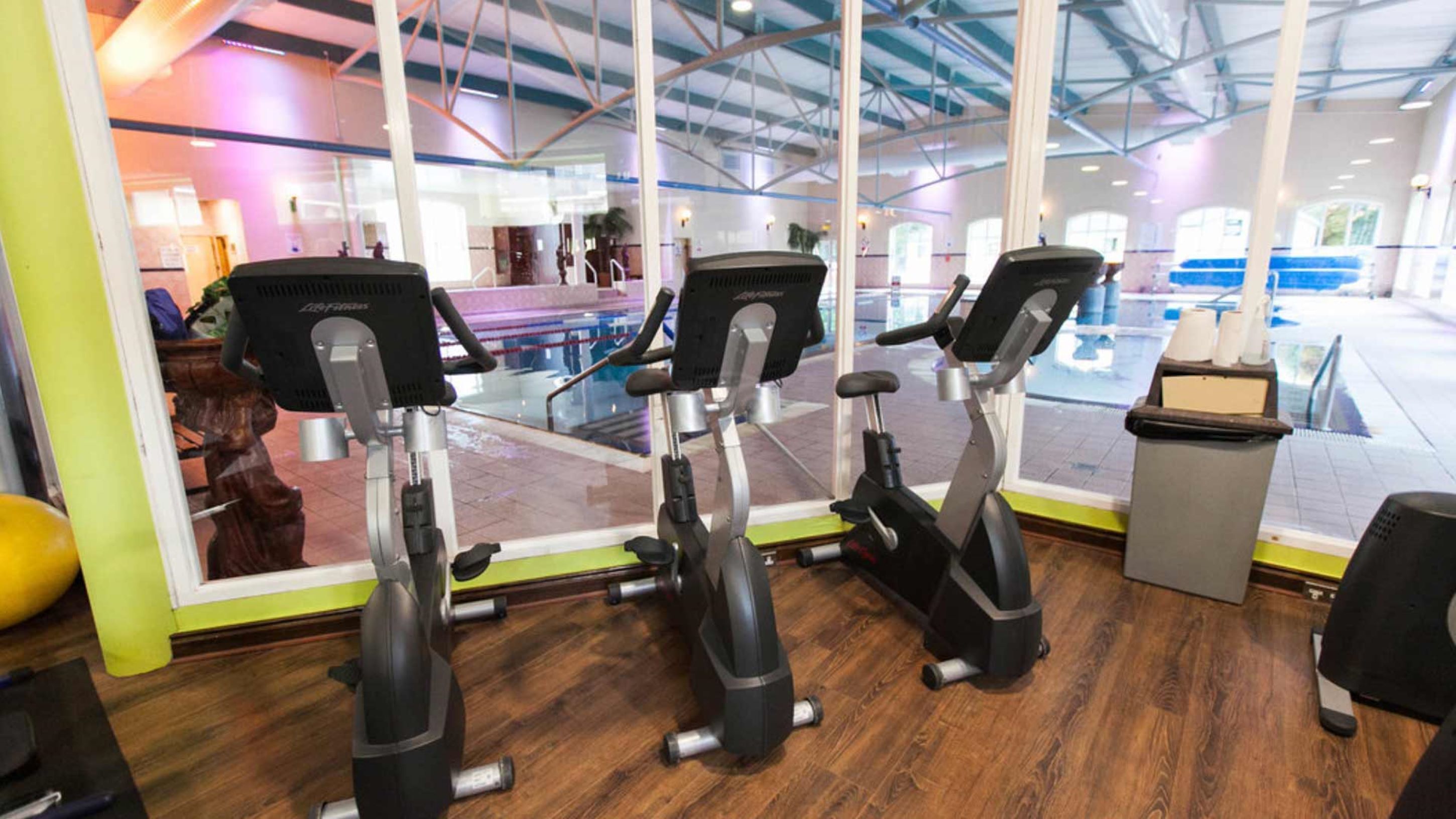 Ginásio interior com três bicicletas ergométricas, piso de madeira e uma grande janela com vista para a área da piscina.