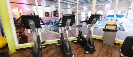 An indoor gym with three stationary bikes, a wooden floor, and a large window overlooking a swimming pool area.