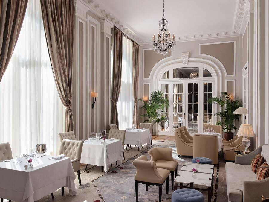 A luxurious hotel lobby with elegant seating, chandeliers, and large windows with curtains.