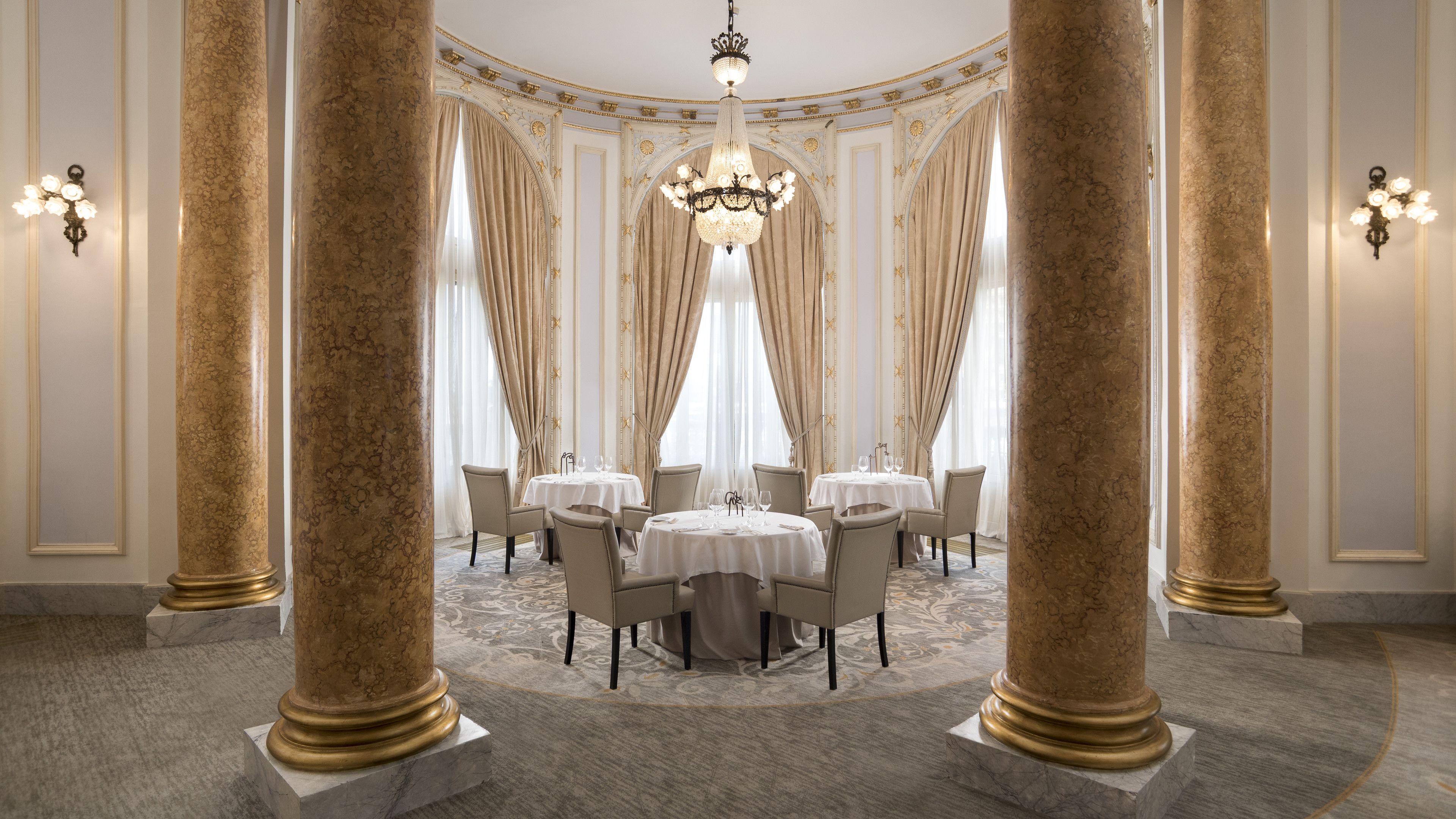 A luxurious dining room with ornate columns, chandeliers, and a central table set for a meal.