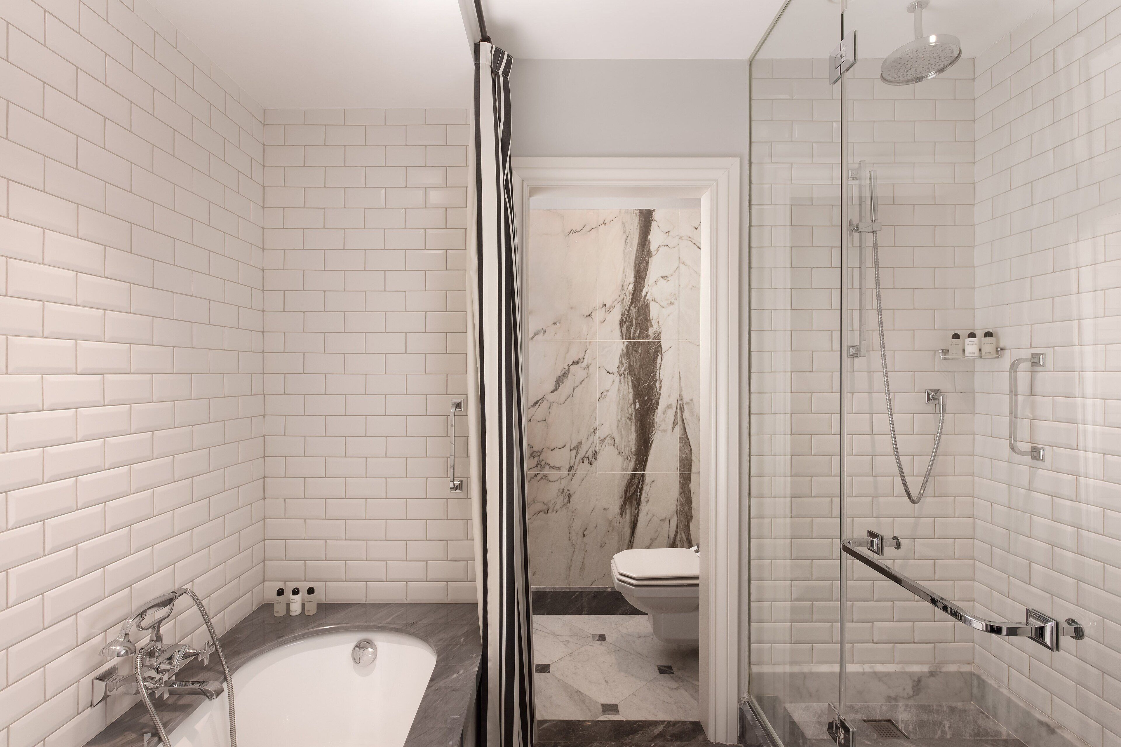 Un baño moderno con azulejos blancos tipo metro, una pared decorativa de mármol, cerramiento de ducha de vidrio y un área de inodoros separada.